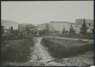 Crowd of people in dark clothing among whitewashed flat-roofed buildings and conifer trees at Marion.