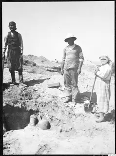 Three people standing at an excavation trench at Marion, with two pottery vessels partly uncovered in the ground in front of them.