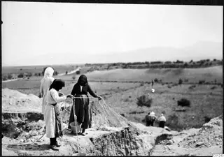 Three women holding a rope at the edge of an excavation trench at Marion, with a bucket on the ground