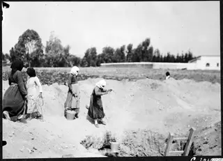 Women in long skirts and head scarves working in an excavation trench at Marion, one lowering a bucket on a rope.