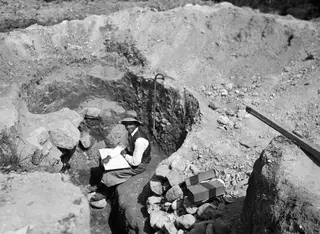 Erik Sjöqvist seated on the edge of an excavated cutting at Marion, resting a drawing board on his knees