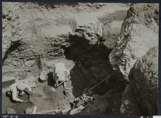 Workers digging in Tomb 37 at Marion, with a bucket, tools and pottery vessels among the soil in the cut.