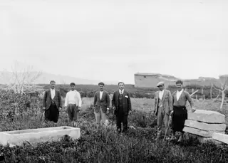 Six men standing in a grassy field at Marion beside a stone sarcophagus and stacked door stones