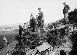 Workers and onlookers standing on a mound of excavated earth at Marion, with low hills and shrubs behind