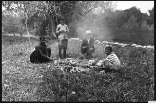 Four people gathered around a small fire on grass near trees at Liso, with smoke drifting across the scene