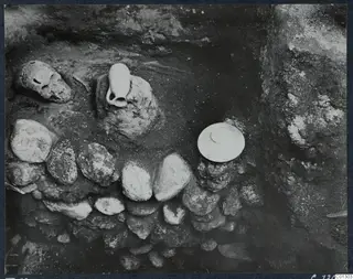 Tomb 59 at Marion during excavation, with a skull and pale vessels among stones and soil.