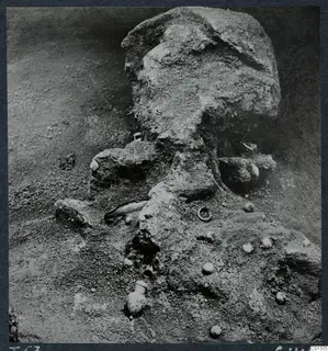 Field photograph of Marion Tomb 57 during excavation, showing a soil-encrusted skull and small finds in place