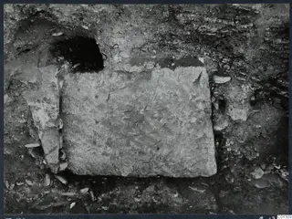Tomb 59 at Marion: a large rectangular stone slab set in excavated soil, with a dark cavity above it and a smaller stone at left.