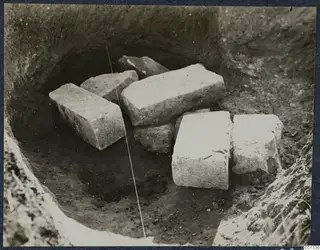 Excavated pit at Marion with several cut stone blocks lying in the soil, Tomb 77