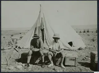 Two men sit on wooden crates outside a large white tent at Marion.