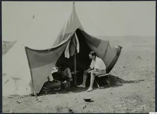 Man seated on a folding stool outside a large canvas tent at Marion, with two men working in the shade inside.