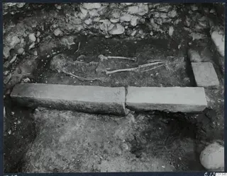Skeleton lying in excavated Tomb 67 at Marion, with two long stone slabs in the foreground.