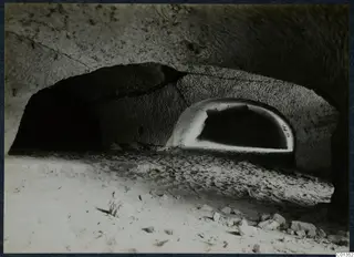Interior of a rock-cut tomb chamber with an arched recess at the back and a sandy floor strewn with stones