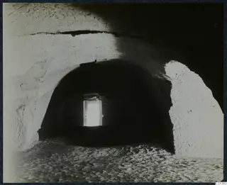Interior of a vaulted rock-cut chamber, its floor strewn with sand and stones, with light from a doorway at the far end.