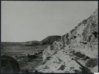 Dirt track running along a rocky cliff edge above a broad valley, with low stone walls on the valley floor