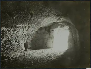Interior of a rock-cut chamber with a bright opening at the back and a sandy floor, Karpas peninsula