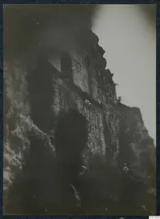 Dark view of a rocky cliff face with a square opening and two small figures on a ledge and at its foot