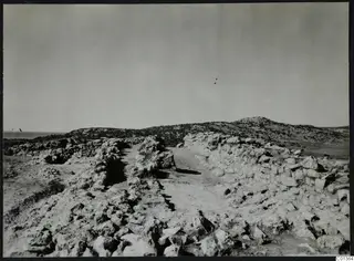 Rough stone walls and a dirt path running through a rocky excavation site toward low hills, with the sea at the far left.