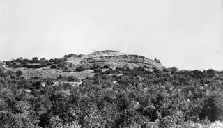 Nitovikla fort on a hill seen from the north-west, with scrub in the foreground and the sea behind.