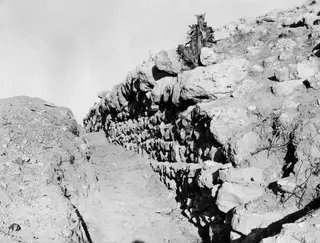 North enclosure wall at Nitovikla, seen from the west, with a long run of stacked stone blocks beside a dirt track.