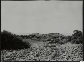 Rocky ground with scrub in the foreground and a low stony mound with a small structure against the sea at Nitovikla