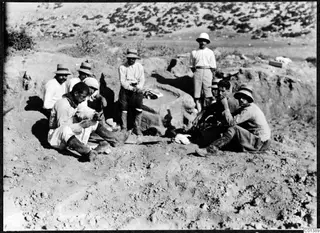 Several men in hats sit and stand beside a large stone block in an excavated area at Oura.
