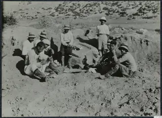 Several men in hats sit and stand on sandy ground beside a large stone block at Oura.