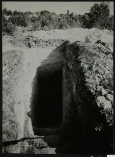 Excavated dromos leading down to a dark tomb entrance at Nitovikla, with stones and scrub behind.