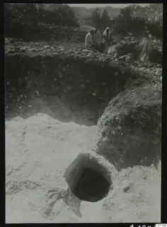 Excavation at Nitovikla on the Karpas Peninsula, with a round opening in pale soil and people seated among stones above