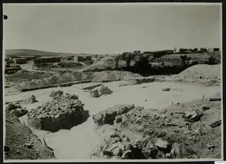Excavated area with exposed stone and mud-brick walls at Ayios Iakovos, with a village of buildings on the hill behind