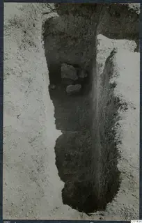 Excavation trench in pale soil with dark cut walls and a few stones lying in a recess, during excavation at Ayios Iakovos.