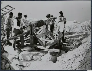 Workers operate a timber winch with a rope drum over a cutting among stone blocks at Vouni, with an animal on the hill behind.