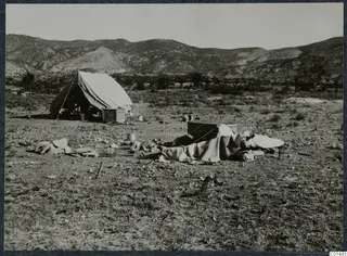 Tent, wooden boxes and draped bedding on bare ground at Ayios Iakovos, with a range of hills behind.
