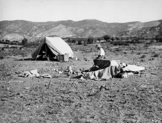 Canvas tent and wooden crates on bare ground at Ayios Iakovos, with a man seated on a crate and hills behind.