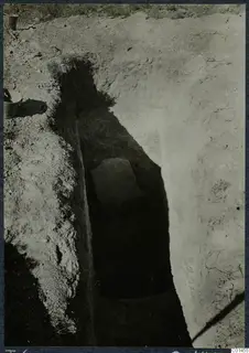 Tomb 6 at Ayios Iakovos during excavation, with a flat stone slab in a deep narrow cut and a bucket at the edge