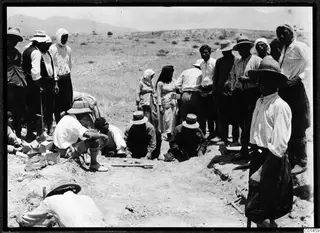 Excavation at Ayios Iakovos with a crowd of people in hats and headscarves working and standing around a shallow trench