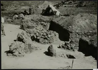 Excavation at Ayios Iakovos with stone walls exposed, a tent and a group of people in the background