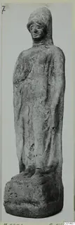 Standing figurine of a robed figure with a hooded head, photographed against a plain grey background