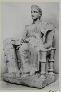 Seated female statue in stone on a throne with turned armrests, shown from the front left.