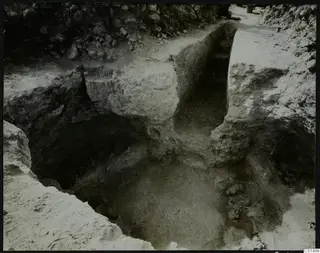 Excavation at Nitovikla showing cut soil faces, a stone wall at the top and small round objects in a dark hollow.