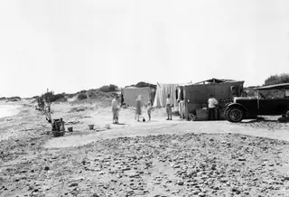 Camp on the beach at Oura with canvas sheets, a matting shelter, several people, a child and an open car.