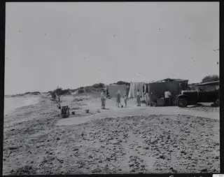 Camp on a stony beach at Oura with people, a child, canvas-screened shelters and an open car.