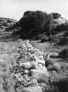 Line of large stone blocks and rubble running across grassy ground at Oura, with bushes and hills behind.