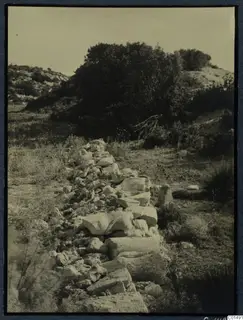 Line of fallen stone blocks and rubble at Oura, with scrub and trees behind on a rise