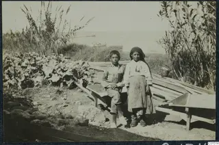 Two children sitting and standing by wooden wheelbarrows on the shore at Soli, with the sea behind them.