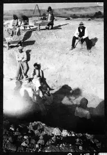 Excavation at Soli with workers in headscarves and a man in a straw hat, a pick, wheelbarrows and stones in the foreground.