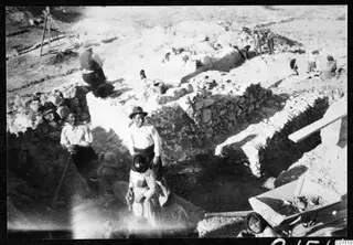Excavation at Soli with rough stone walls, standing workers in the foreground and a wheelbarrow at lower right.