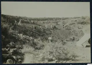 Workers with picks and shovels excavating a sloping hillside at Soli, with a stone wall above and a tent on the right.