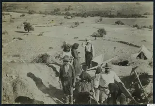 Workers digging an excavation trench at Soli, with a white tent and a wheelbarrow in an open, sparsely wooded landscape.