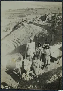 Work group of an older man, young men and women posed on an excavation mound at Soli, with tools and stone walls behind.